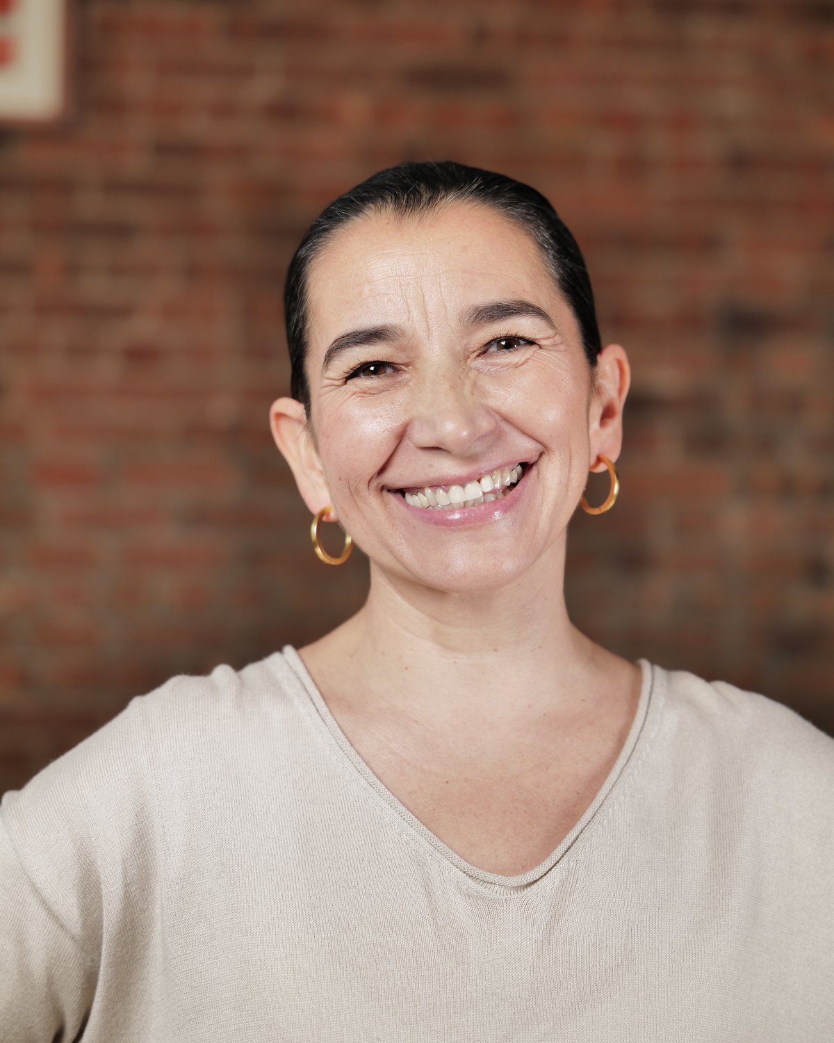 Alejandra Belmar — close portrait, direct gaze, warm smile, brick wall background.