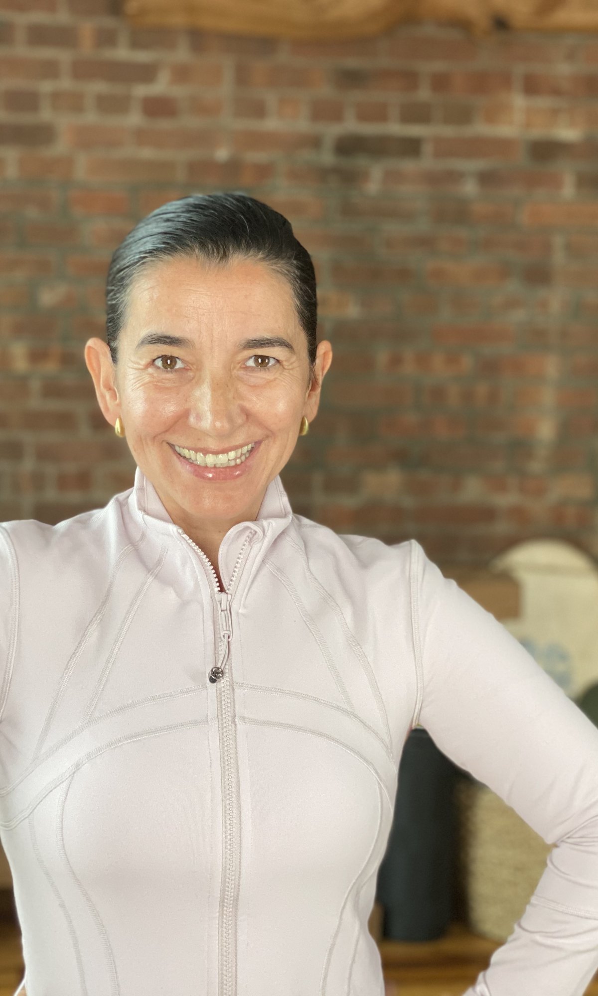 Alejandra Belmar — warm full smile, hands on hips, lavender activewear against a brick wall in her Brooklyn studio.