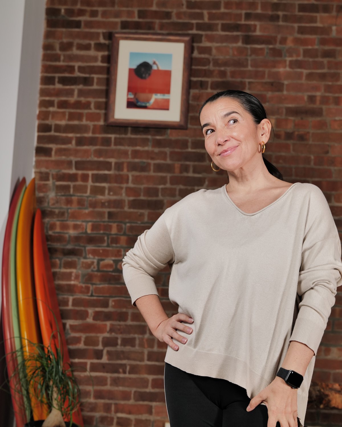 Alejandra Belmar — portrait against a brick wall, cream sweater, hand on hip, looking off-camera.