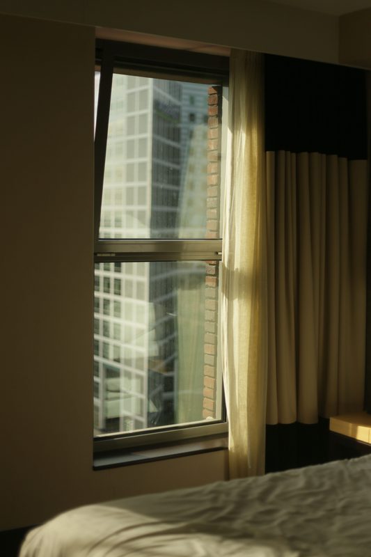Early morning light coming through a bedroom window onto the corner of a made bed, the city quiet outside.