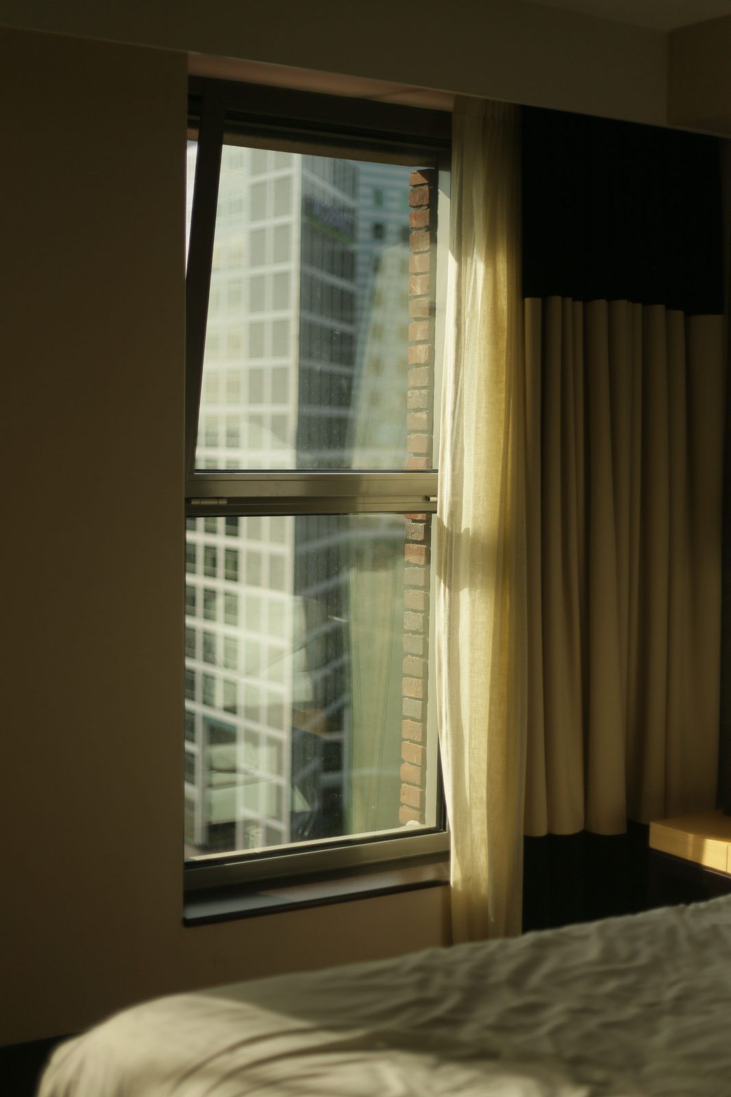 Early morning light coming through a bedroom window onto the corner of a made bed, the city quiet outside.