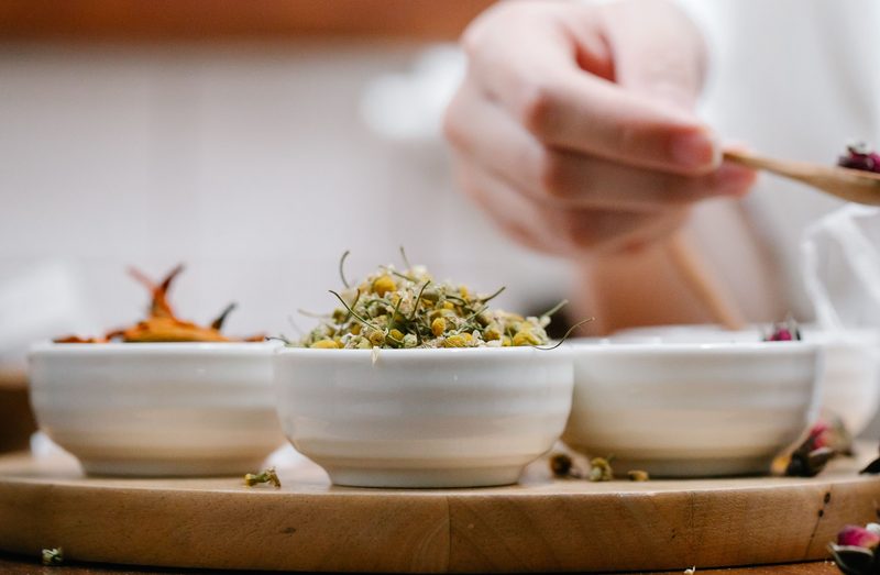 Small white bowls of dried chamomile and flower petals on a wooden board, a hand spooning dried rosebuds in the background.