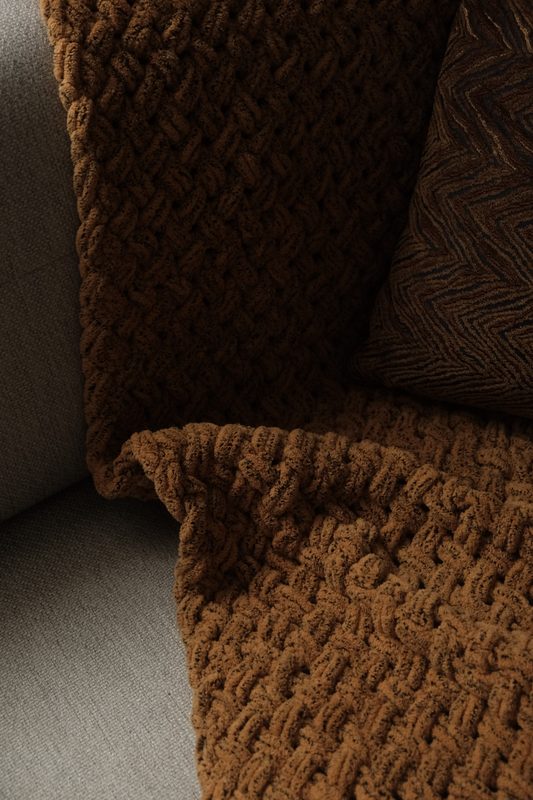 Close-up of a chunky hand-knit blanket in warm brown tones draped over a gray sofa.