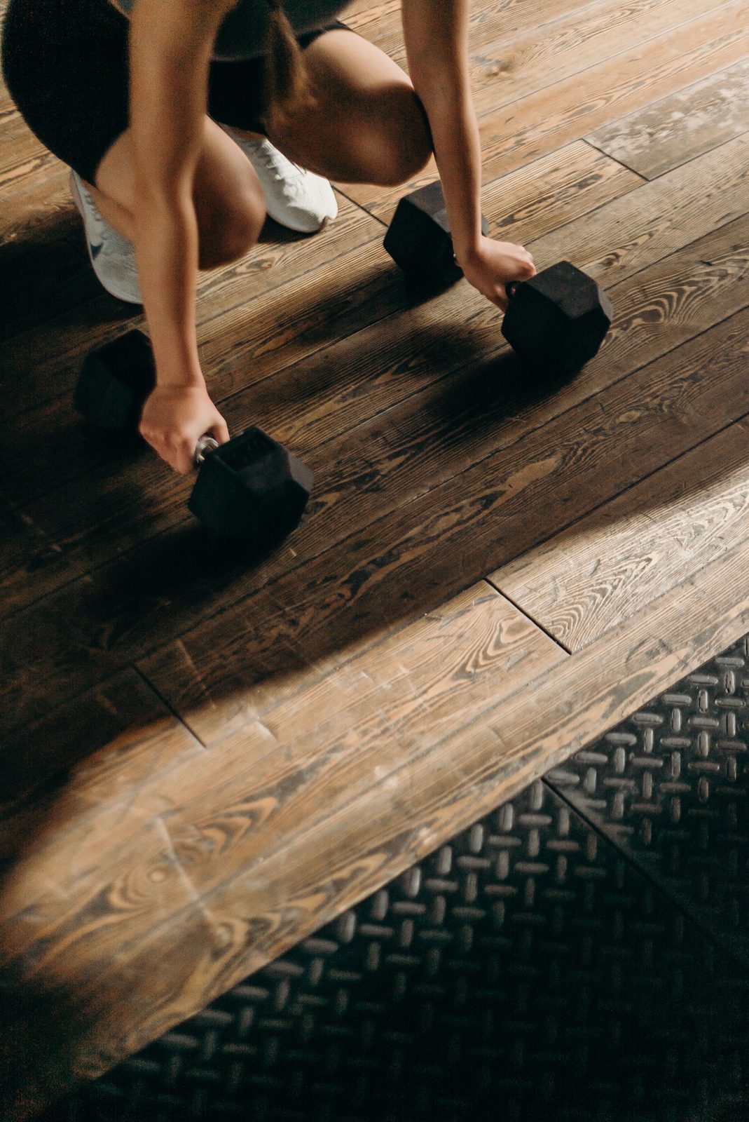 A woman crouches on a wooden floor, gripping two hex dumbbells before lifting.