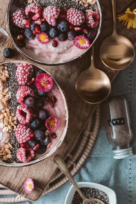 Two bowls of berry smoothie topped with frozen raspberries, blueberries, pomegranate, chia seeds and oats, with brass spoons and a jar of flax on a wooden board.