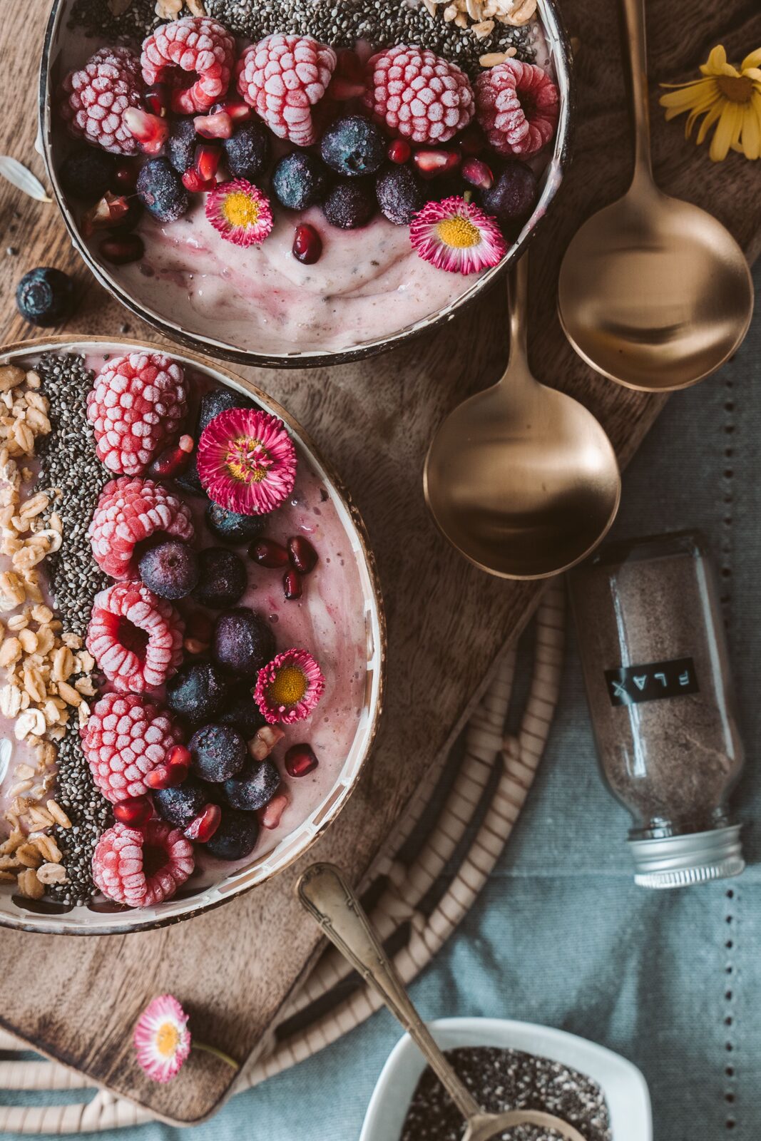 Two bowls of berry smoothie topped with frozen raspberries, blueberries, pomegranate, chia seeds and oats, with brass spoons and a jar of flax on a wooden board.