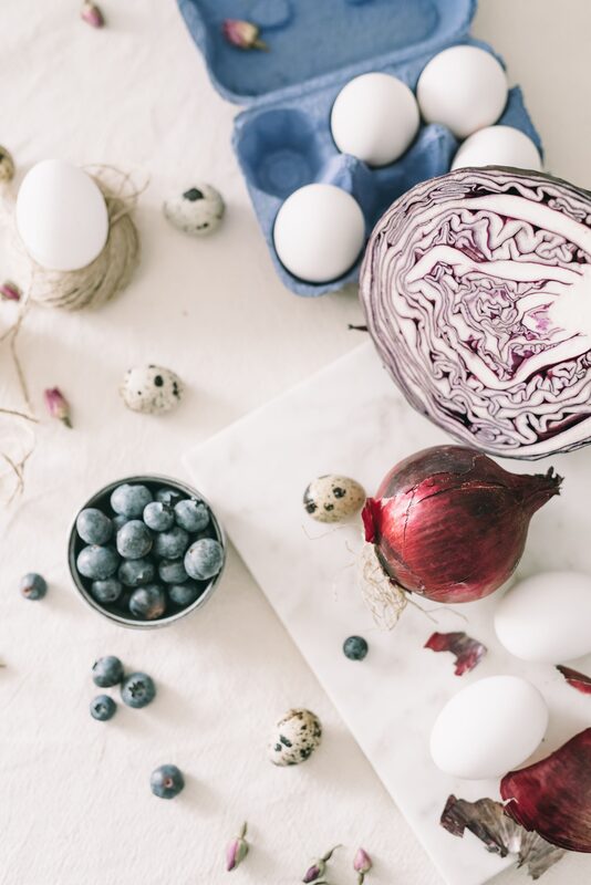 A halved red cabbage, a red onion, a bowl of blueberries, and white and quail eggs on a marble board over pale linen.