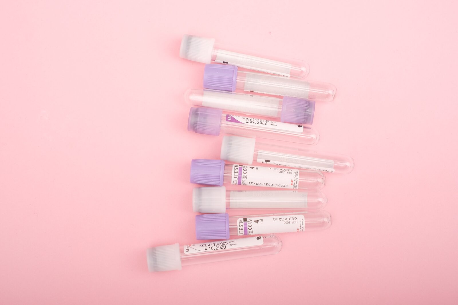 Clear laboratory collection tubes with lilac and white caps, scattered on a pale pink surface.
