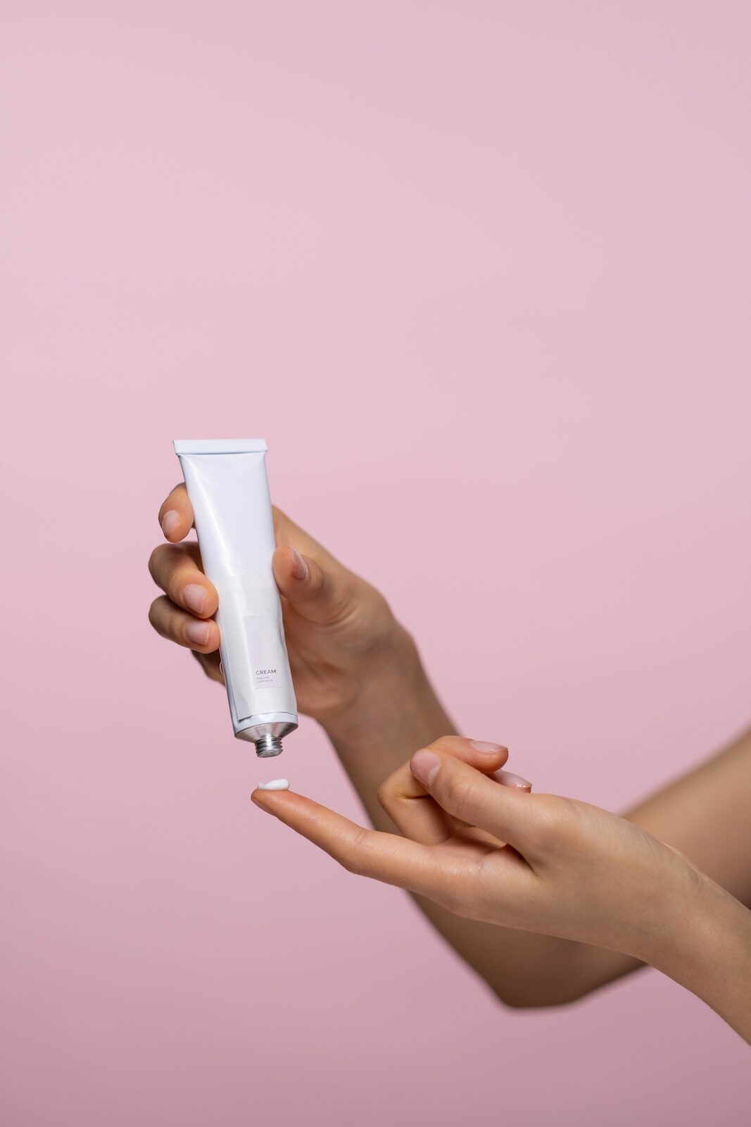 Two hands against a soft pink background, one holding a plain white tube, the other receiving a small amount of cream on a fingertip.