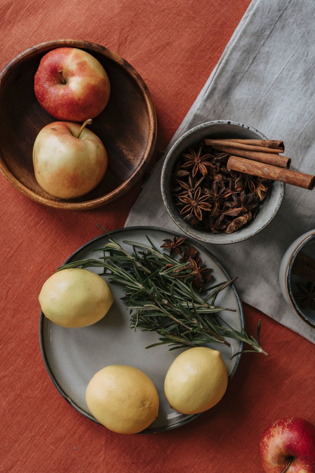 Apples in a wooden bowl, lemons and rosemary on a stoneware plate, and star anise with cinnamon on rust-colored linen.