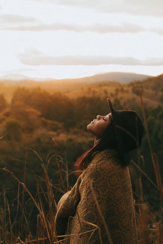 A woman in a wide-brimmed hat and woven coat, face lifted to the sky, golden hills behind her.
