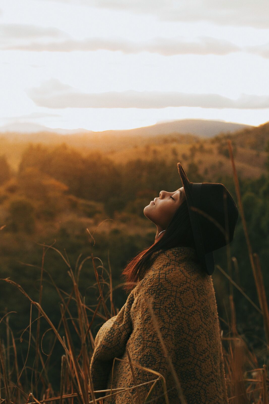 A woman in a wide-brimmed hat and woven coat, face lifted to the sky, golden hills behind her.