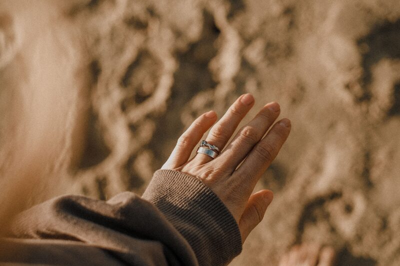 A hand resting open on warm sand, wearing two slim rings, the cuff of a soft knit sleeve at the wrist.