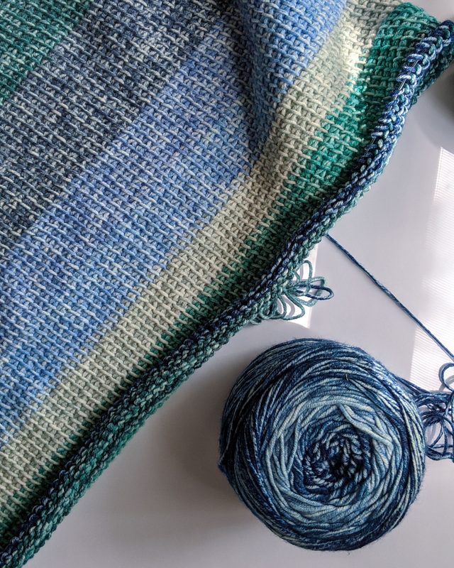 A crocheted blanket in bands of blue, cream, and green in progress, beside a ball of blue variegated yarn in soft window light.