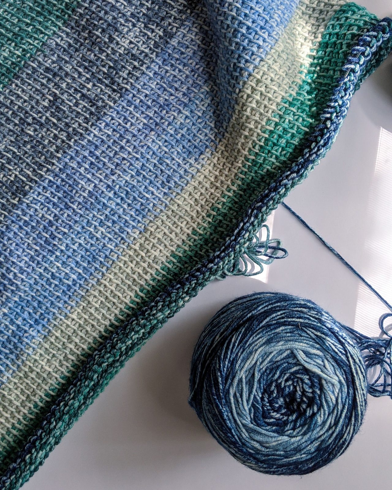 A crocheted blanket in bands of blue, cream, and green in progress, beside a ball of blue variegated yarn in soft window light.