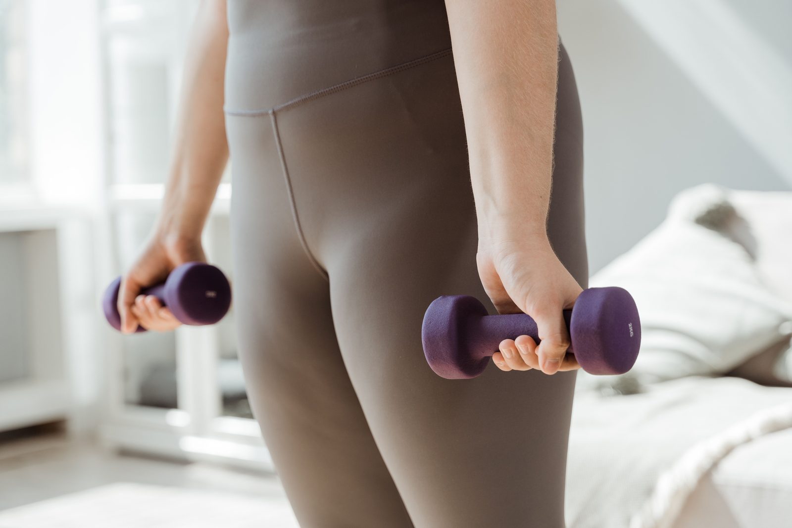 A woman in taupe leggings stands in a bright room holding a small purple dumbbell in each hand.