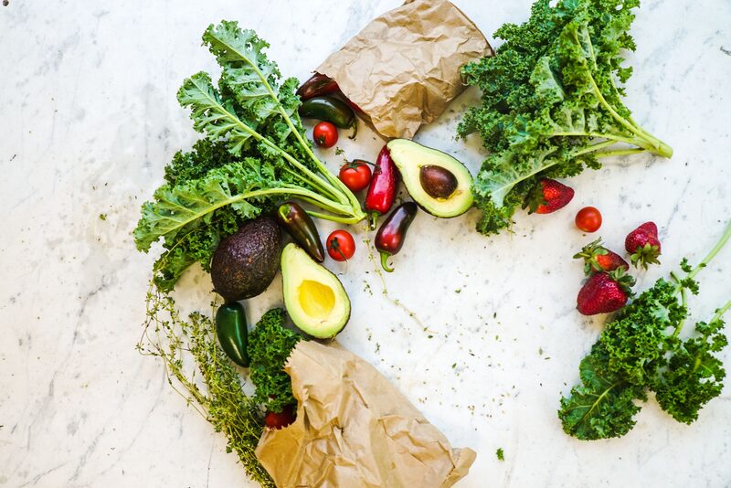 Kale, avocado halves, cherry tomatoes, chilies, strawberries and thyme spread across a white marble surface with brown paper bags.