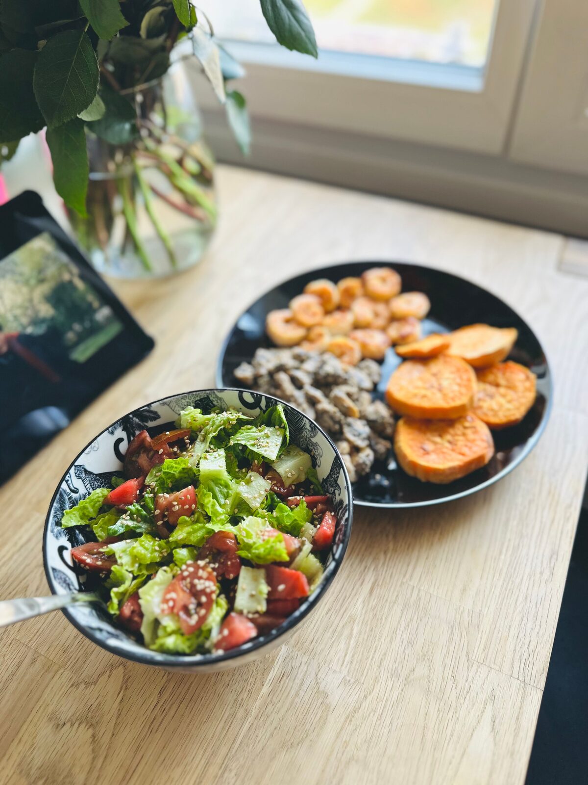 A bowl of tomato and romaine salad with sesame beside a plate of roasted sweet potato and plantain, on a wooden table by a window.