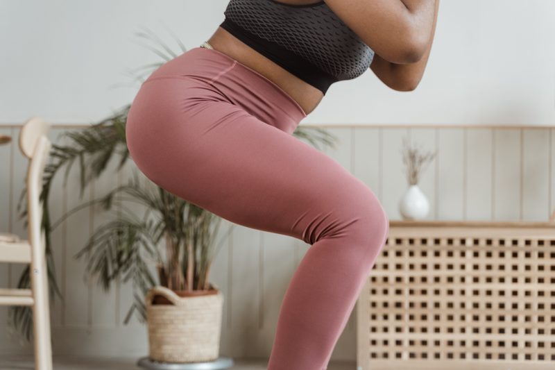 A woman in rose-colored leggings lowers into a squat in a bright room with a plant and a wooden chair.