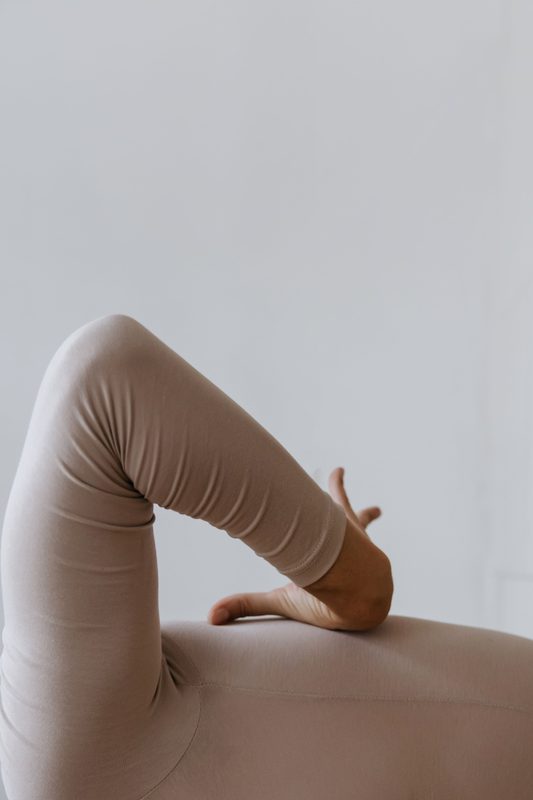 Close-up of a bent leg in soft taupe leggings, a hand resting on the thigh, against a pale wall.