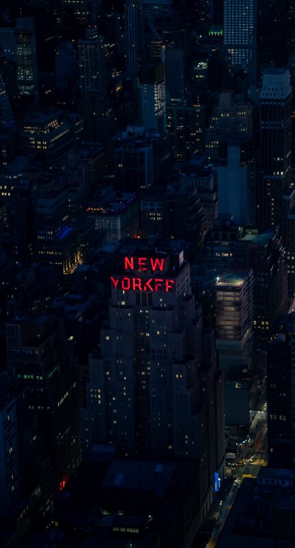A city at night seen from above, buildings dark and lit windows scattered, one red rooftop sign glowing.