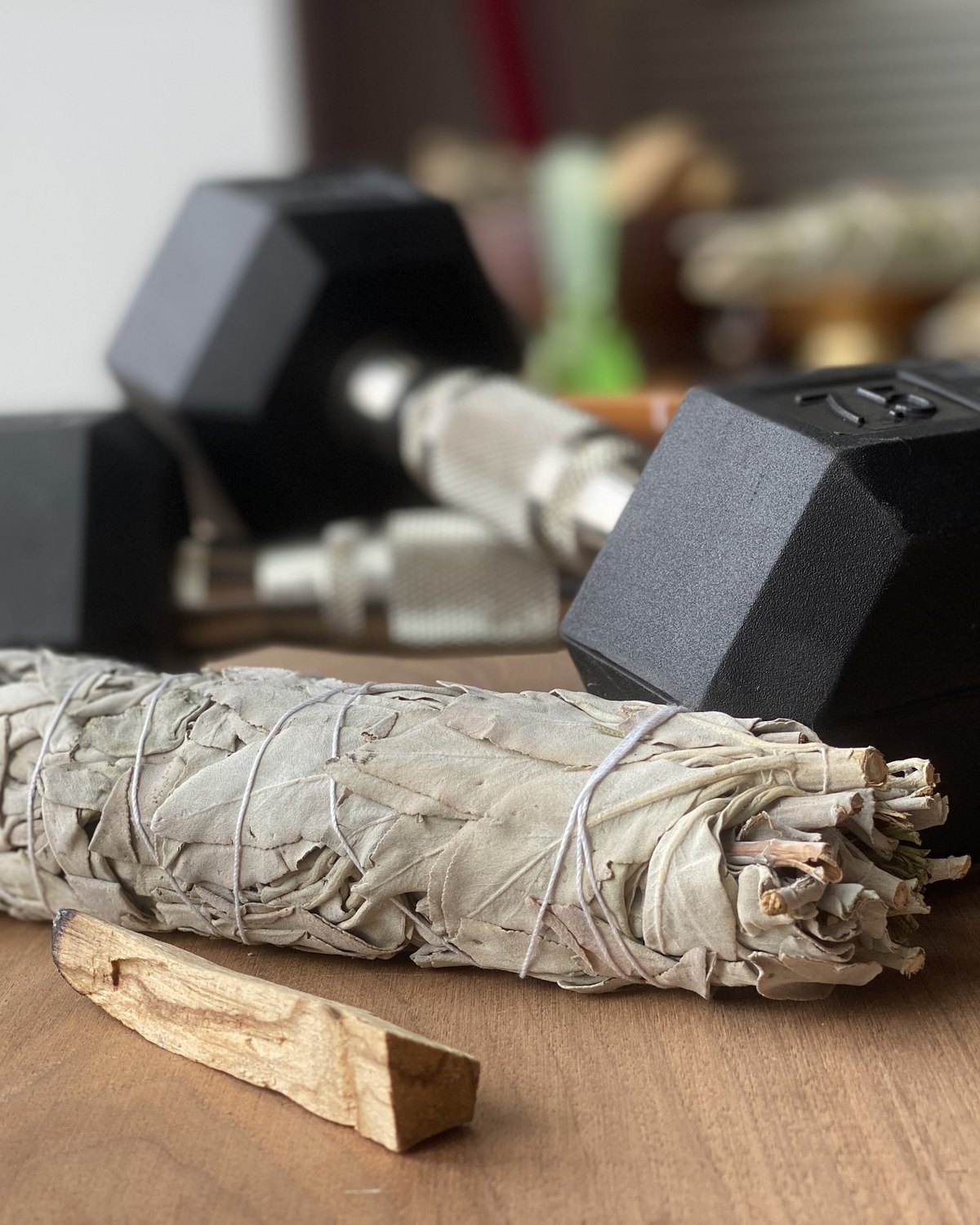 Two black hex dumbbells on a wooden surface with a white sage bundle and palo santo in the foreground — natural light, shallow depth of field.